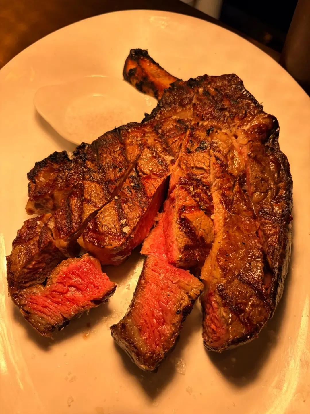 Sliced bone-in côte de boeuf on a white plate, charred outside and rosy pink inside, beside a small dish of flaky salt
