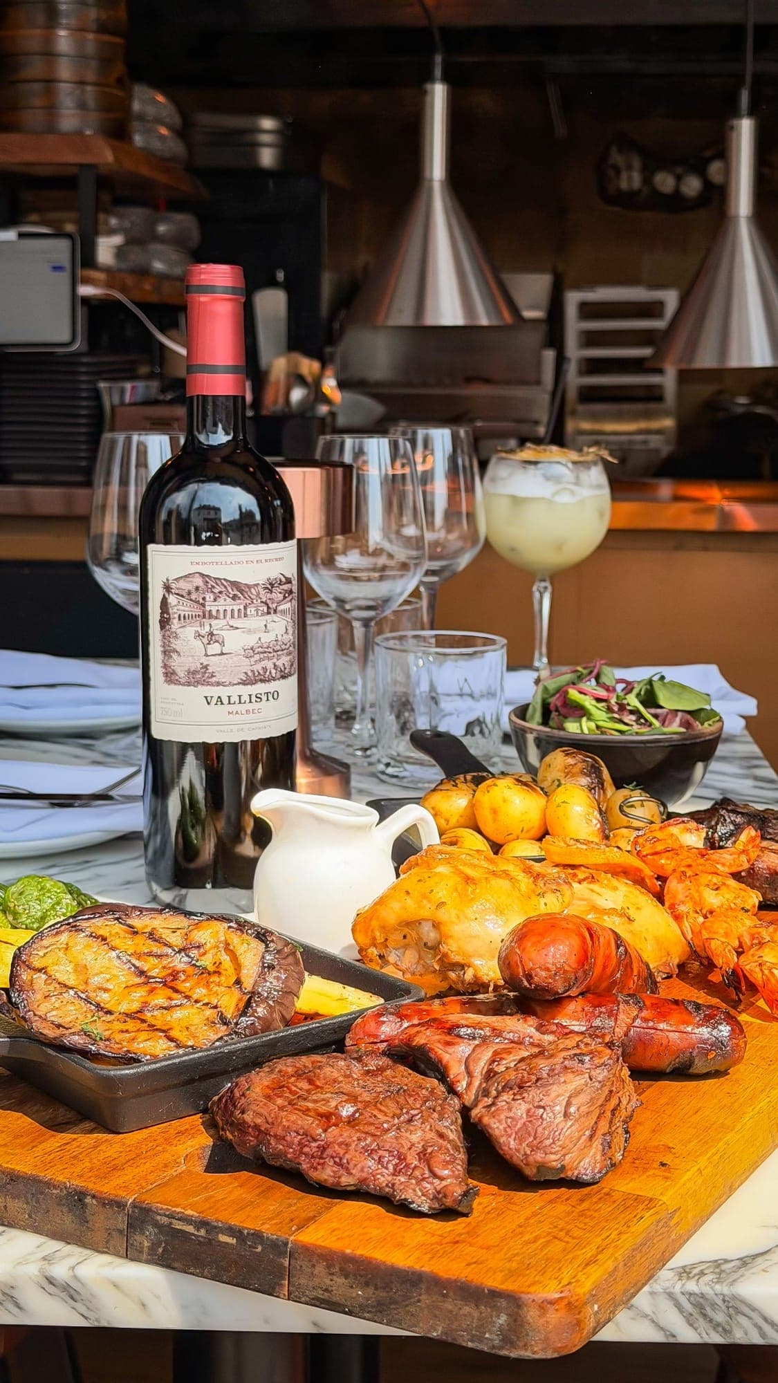 A wooden board of grilled steaks, chicken, chorizo and prawns with roast potatoes, laid for sharing beside a bottle of Malbec