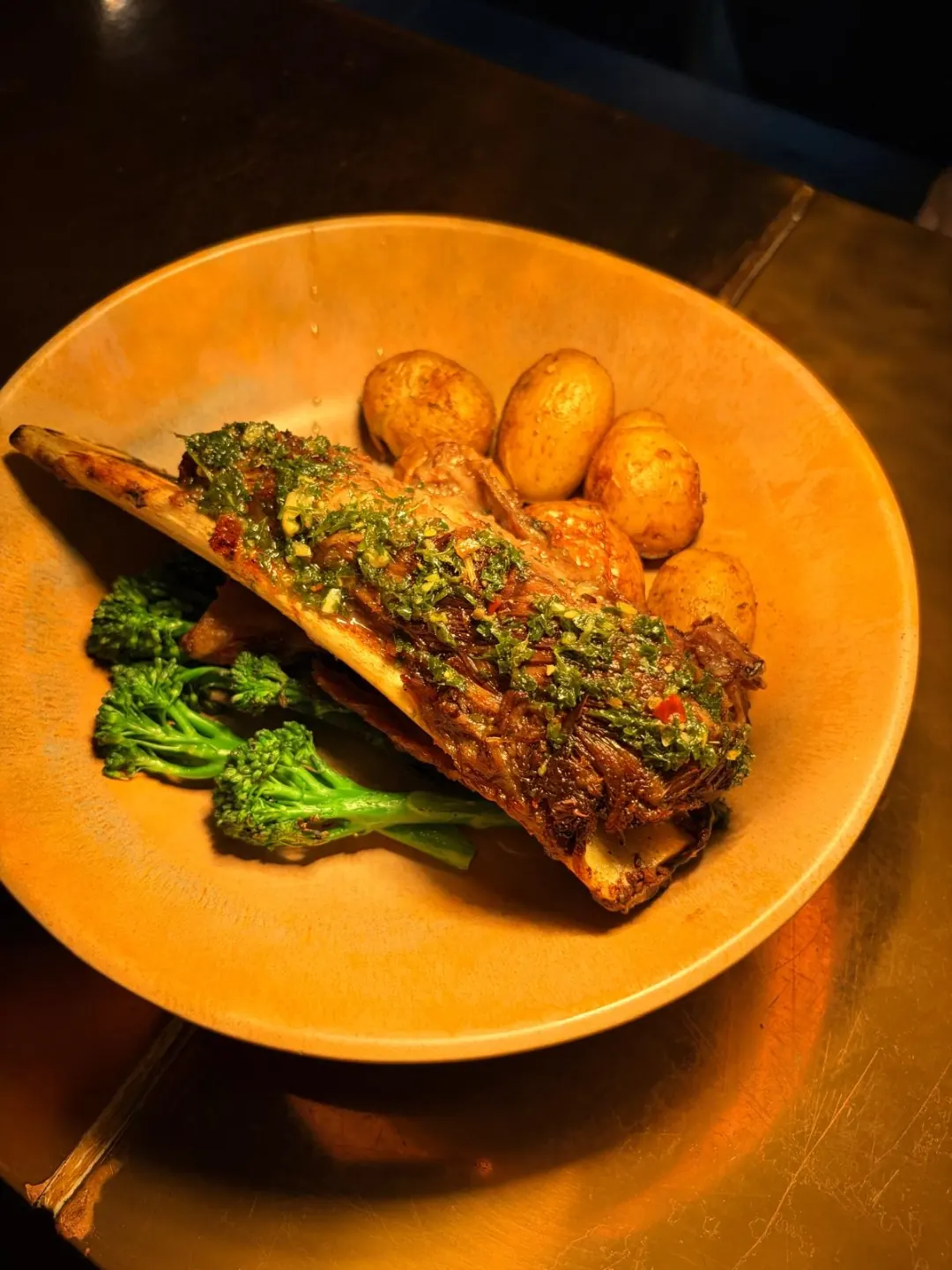 Fire braised short rib on the bone, glossed with green chimichurri, beside charred broccolini and roast baby potatoes in a terracotta bowl