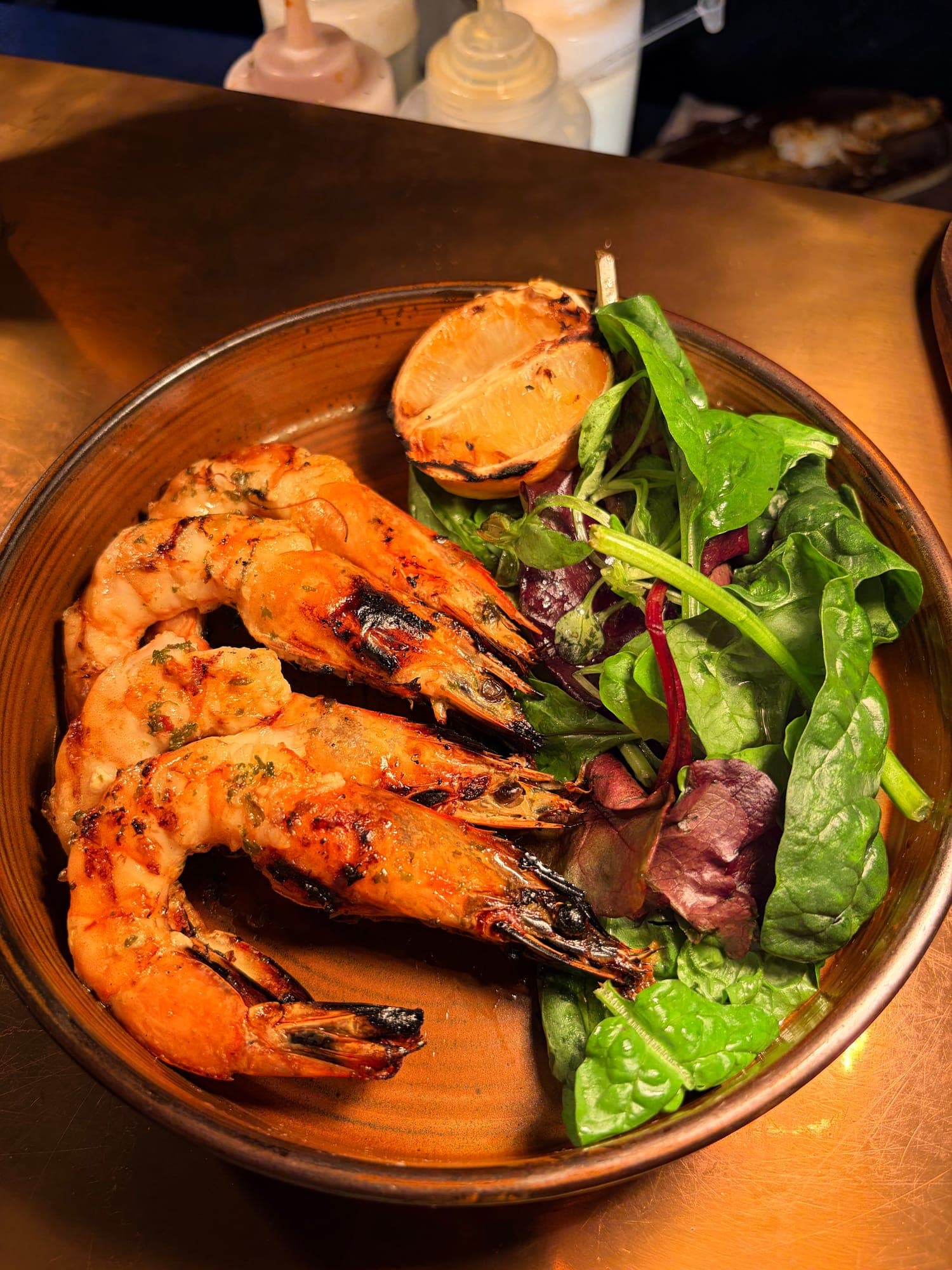 Four grilled king prawns glossed with garlic butter and herbs in a terracotta bowl with a charred lemon half and salad leaves