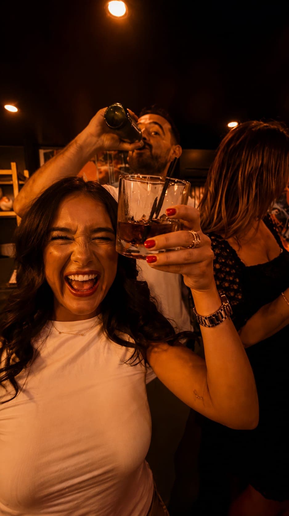 A guest laughing and raising a glass over ice while a friend tips back a bottle behind her, in the warm low light of NORA
