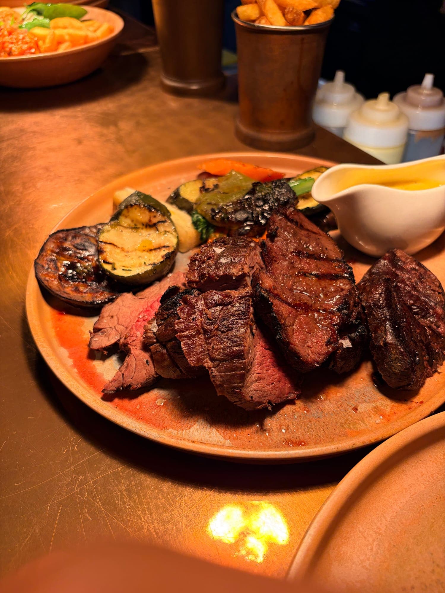 Sliced fire-grilled steak with charred aubergine and courgette, a jug of sauce and a cup of chips on a terracotta plate