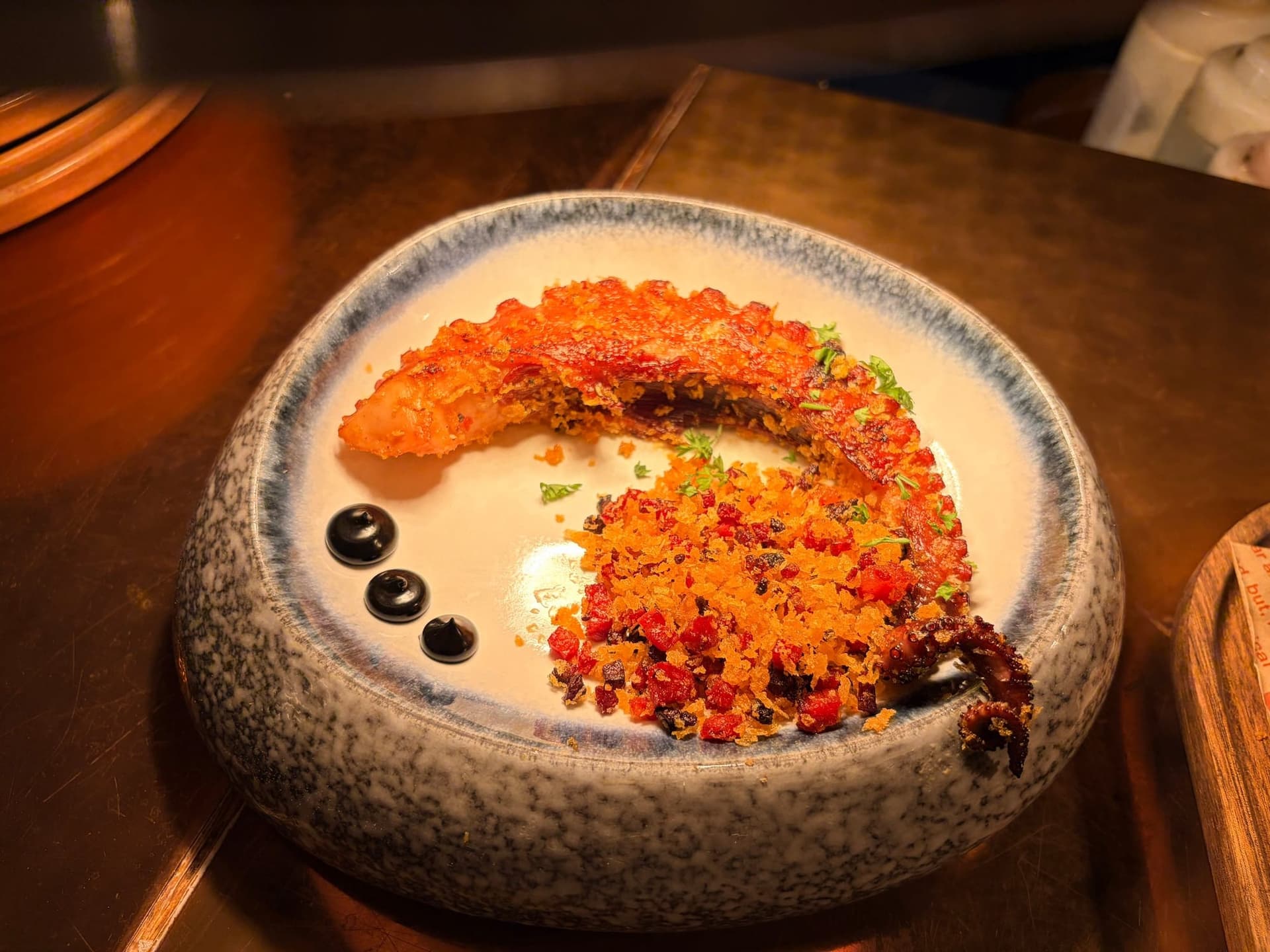 Charred octopus tentacle in a speckled bowl with crisp chorizo crumb and three dots of black mayo