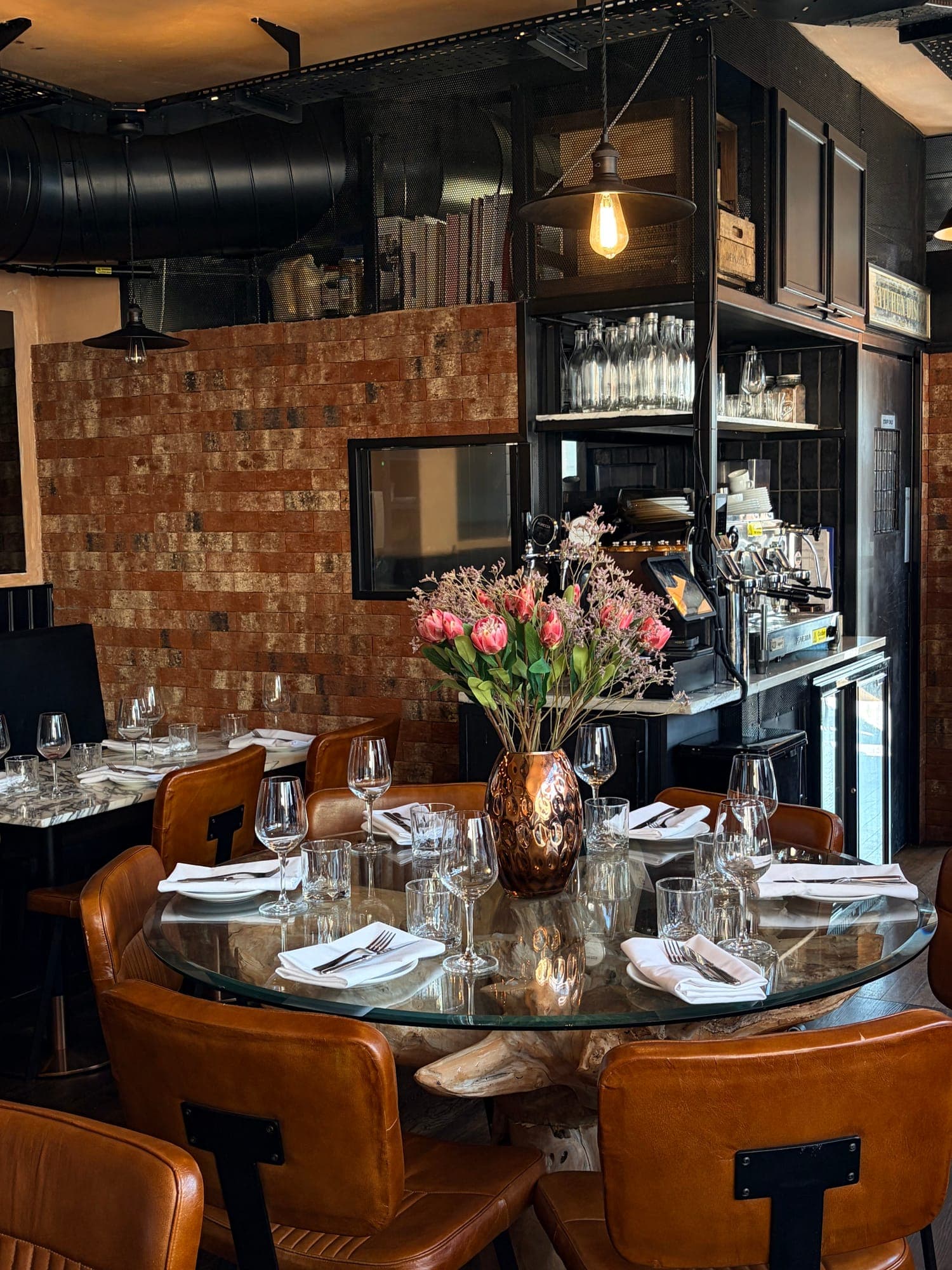A round glass-topped table set with wine glasses and folded white napkins, a copper vase of pink flowers, and tan leather chairs